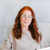 A woman with long red hair wearing glasses and a beige top sits on a white chair.